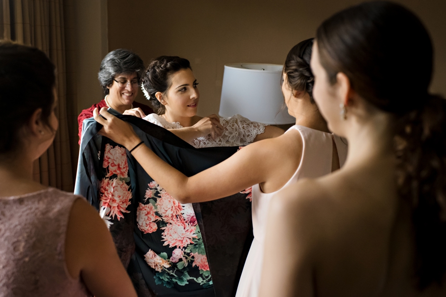 The bridesmaids block the view of the brides mother helping her in her wedding dress.